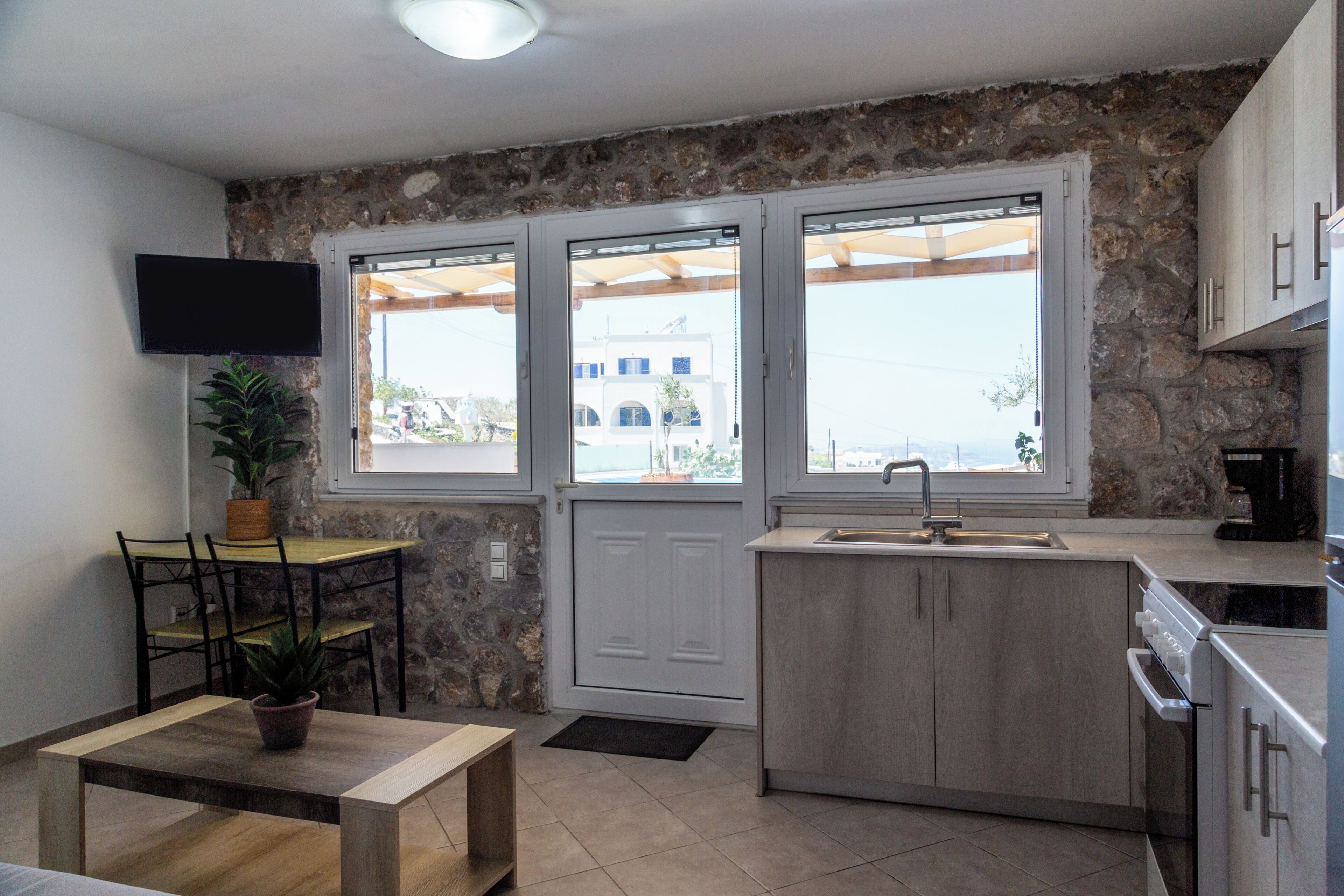 A compact kitchen with stone walls, a dining area with a table and chairs, a TV mounted on the wall, and a view through the windows.