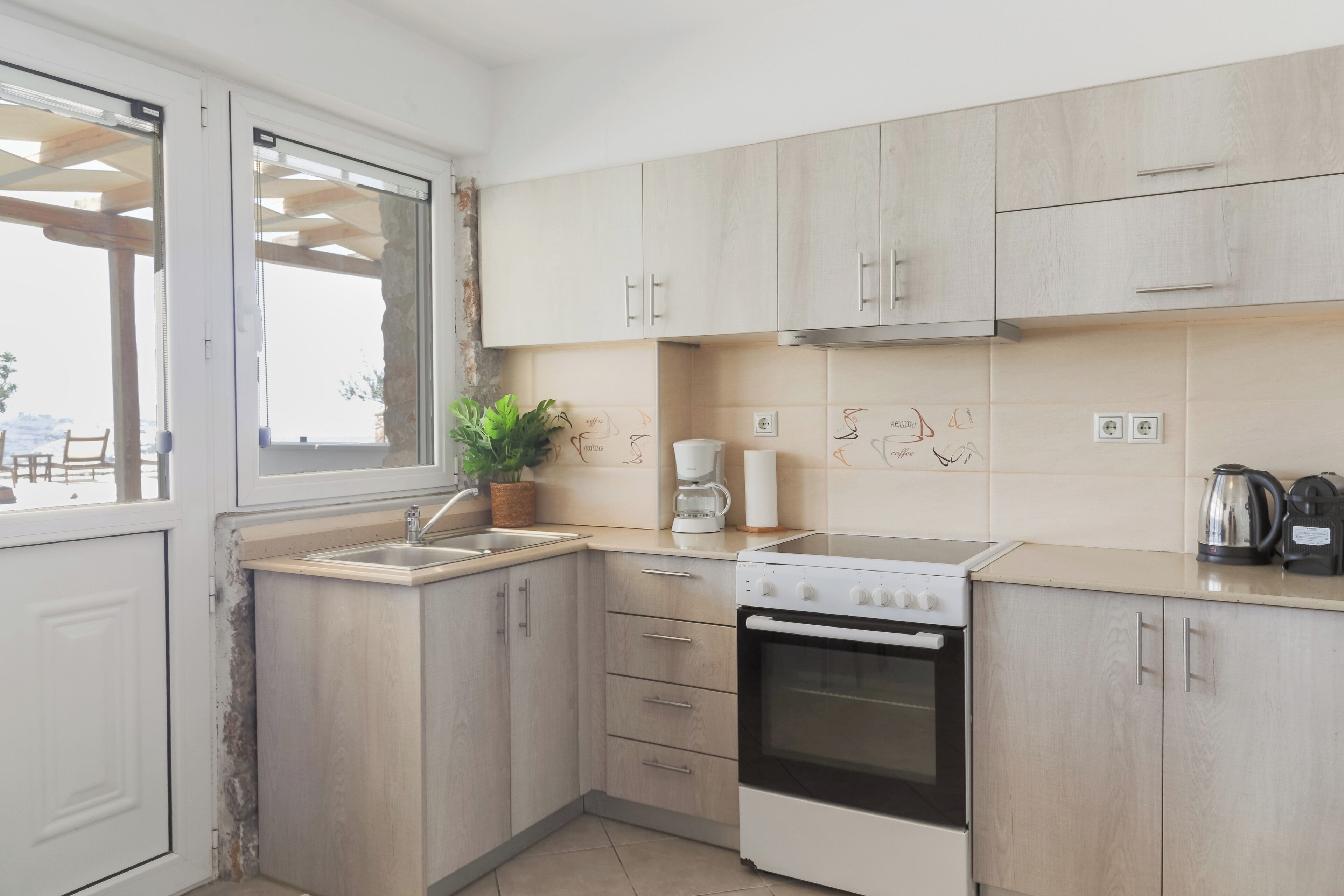 A modern kitchen with light wood cabinets, a built-in oven, a coffee maker, and a potted plant.