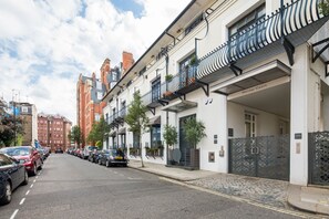 A row of modern buildings with balconies, a cobblestone street, and parked cars.
