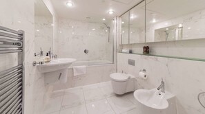A modern bathroom with a bathtub, toilet, and sink, marble walls, and a glass shelf.