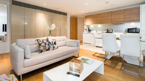 Un salon moderne avec un canapé, une table basse et des fauteuils. On aperçoit une cuisine à l’arrière-plan, avec des meubles blancs et un four à micro-ondes.