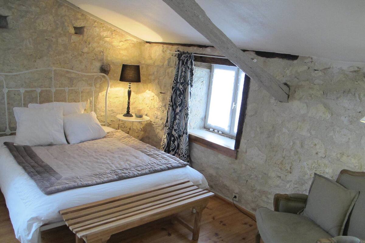 Een slaapkamer met een stenen muur, een bed met witte beddengoed, een bedlampje, een raam met gordijnen en een houten bankje.