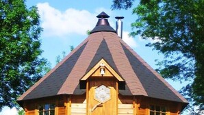 Une cabane en bois avec un toit conique, une porte centrale ornée d’une fenêtre en forme de losange et une cheminée.