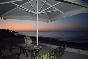 Ein Sitzbereich im Freien mit Tisch und Stühlen, einem Sonnenschirm und Blick auf das Meer bei Sonnenuntergang.