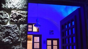 Un pasillo tenuemente iluminado con paredes de piedra y un tono azulado, que conduce a una habitación con una puerta y una ventana.