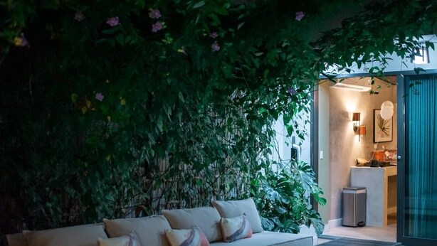 A cozy living area with a sofa and a coffee table, surrounded by lush green foliage and a glass door leading to a kitchen. - Trópica Beach Hotel (San Juan)
