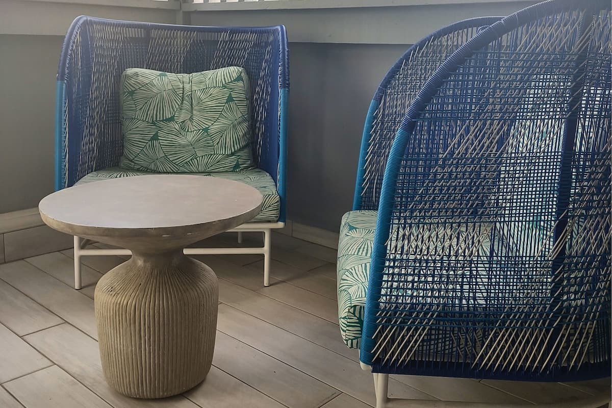 A room with two wicker chairs, one with a green patterned cushion and the other with a blue patterned cushion, and a round table with a ribbed base.