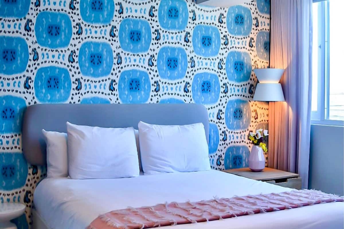 A neatly made bed with white linens and a pink throw blanket, a bedside table with a lamp, and a patterned wall in the background.