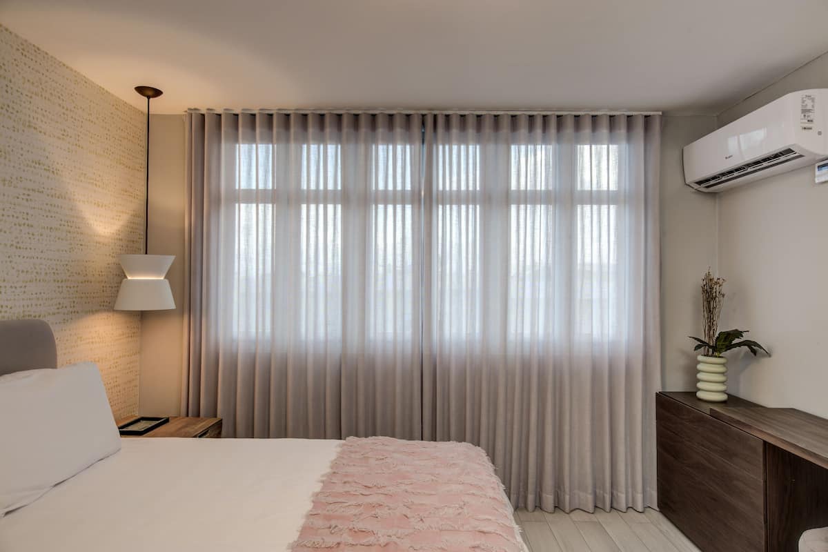 A hotel room with a bed, a nightstand, a lamp, a window with curtains, an air conditioning unit, and a small plant on a console table.