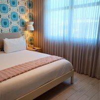 Una cama bien hecha con una colcha blanca y un plaid rosa, una lámpara de noche y una pared con diseño azul y blanco.
