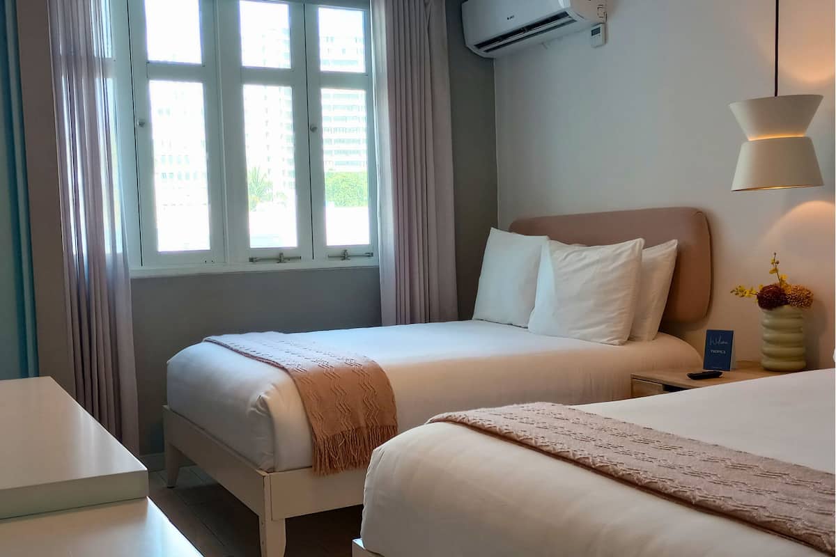 A hotel room with two beds, a window with curtains, an air conditioning unit, a bedside table with a lamp, and a vase with flowers.