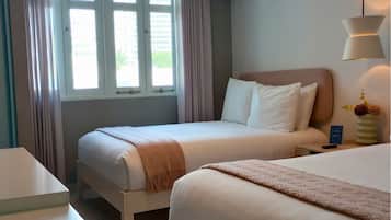 A hotel room with two beds, a window with curtains, an air conditioning unit, a bedside table with a lamp, and a vase with flowers.