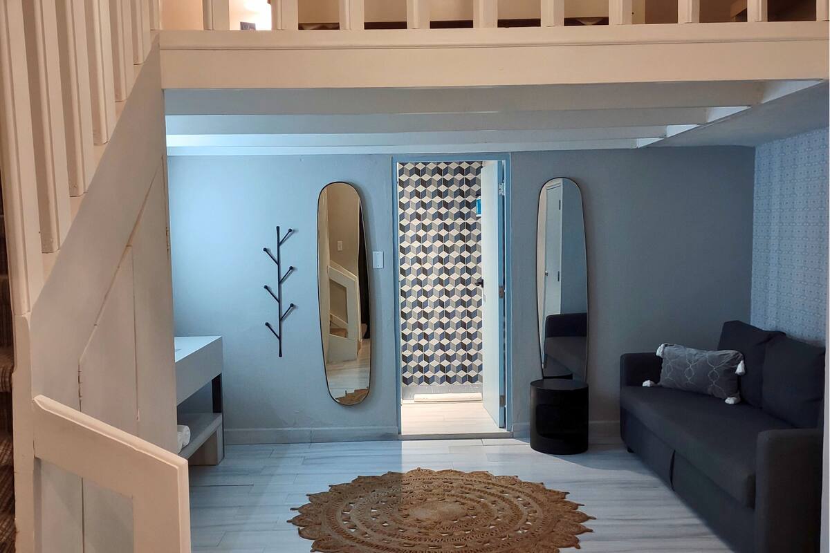 A modern living room with a staircase, a large mirror, a patterned wall, and a dark sofa.