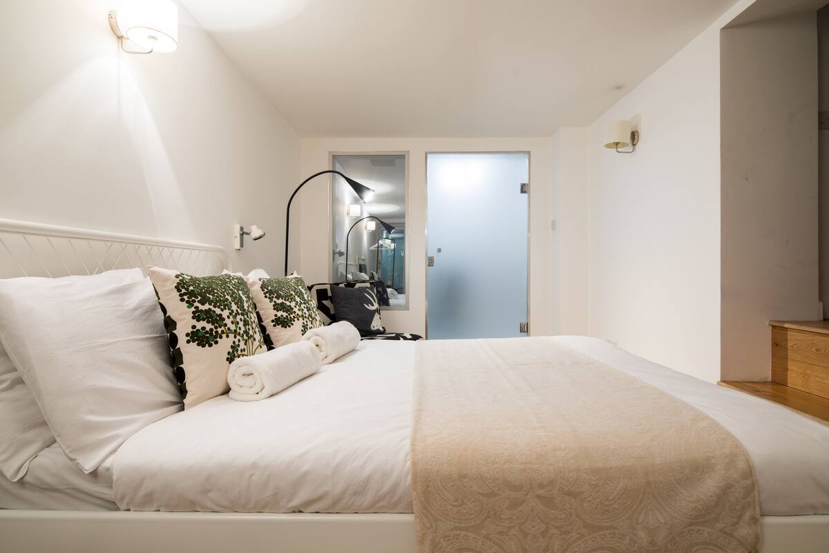 Una cama bien tendida con sábanas blancas y una manta color beige, una lámpara de noche y una lámpara de pared.