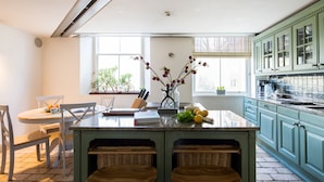 A kitchen with a central island, two wicker stools, a dining table with chairs, and a window with plants. - One Palace Gate (London)