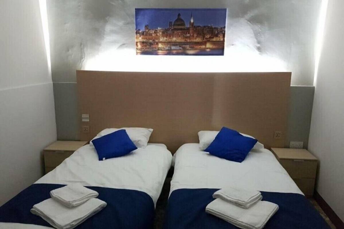 Habitación de hotel con dos camas, cada una con una almohada azul y ropa de cama blanca. Encima de las camas, hay un cuadro con una vista de la ciudad.