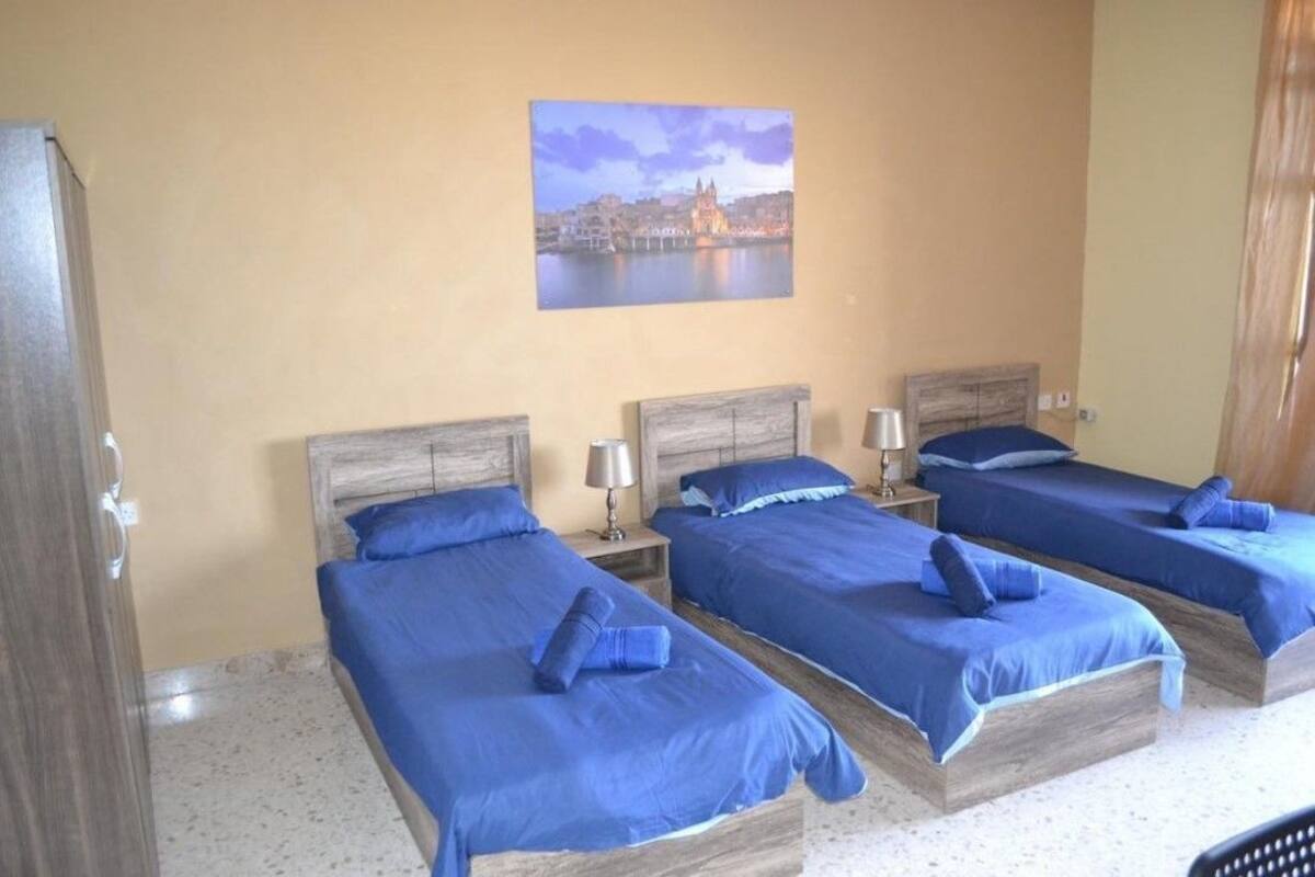 Una habitación de hotel con tres camas, cada una con ropa de cama azul y cabeceros de madera. En la pared sobre las camas hay un cuadro de un paisaje urbano.