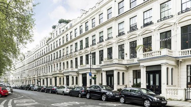 A row of white townhouses with uniform architecture, parked cars along the street, and a clear sky. - Cleveland Square Mansions (London)