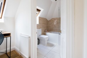 A modern bathroom with a white toilet, a bathtub, and a shower. There is a radiator on the wall and a small table with a chair. - Cornwall Gardens by Lime Street (London)