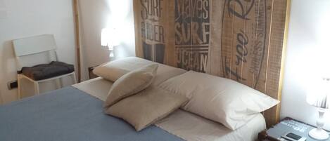 Un dormitorio con una cama grande, cabecera de madera con texto relacionado con el surf, una mesita de noche pequeña y un tocador con lámpara.