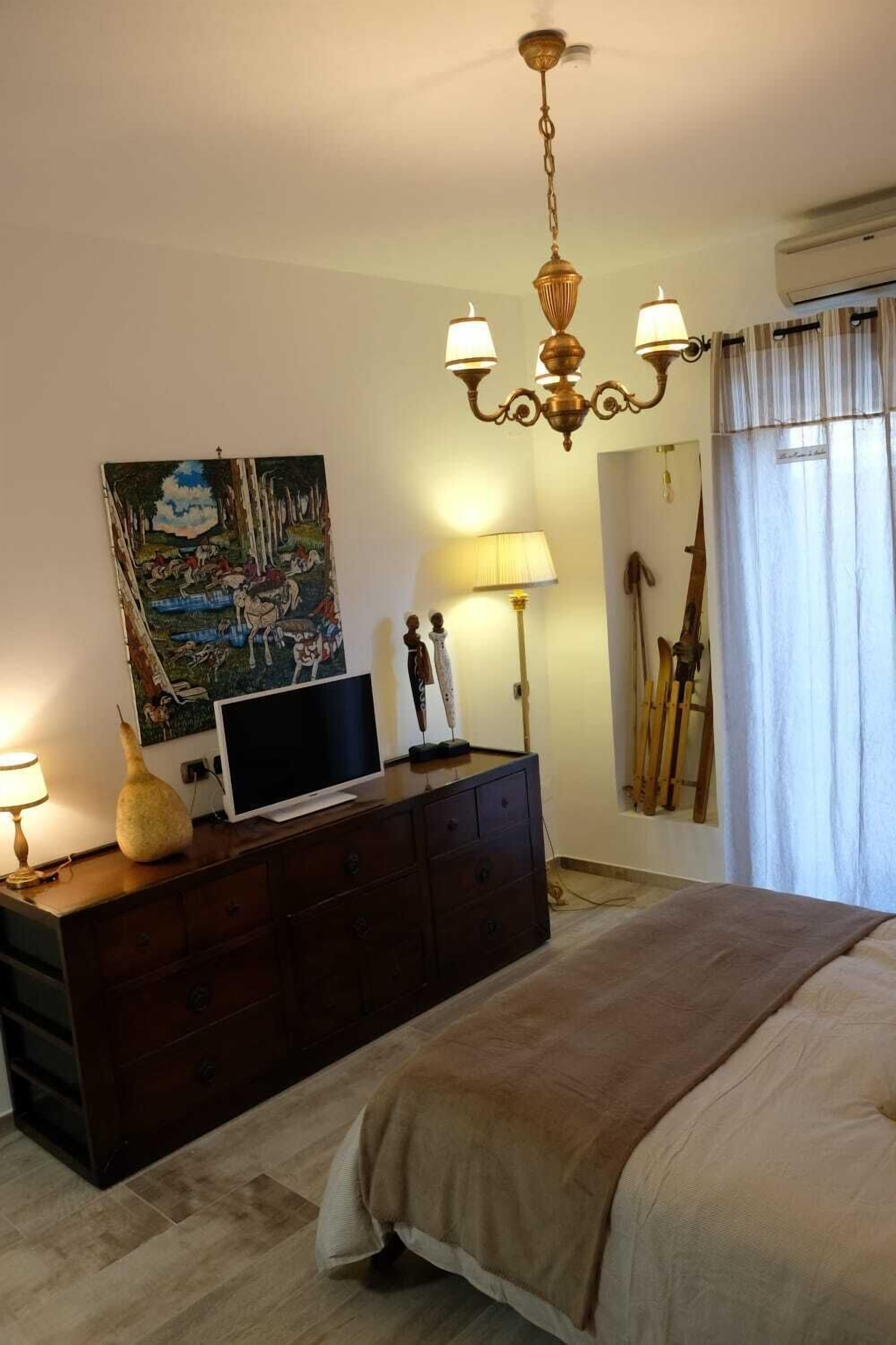 Un dormitori amb un llit, una petita arca, un quadre, una làmpada i esquís recolzats a la paret.
