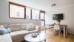 A modern living room with a sofa, a dining table with chairs, a TV mounted on the wall, and large windows with blinds. - Richardsons Mews by Lime Street (London)