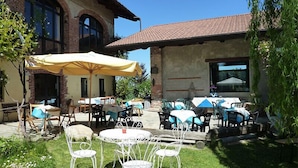 An outdoor patio area with tables and chairs, some covered by umbrellas, and a building with a tiled roof in the background. - Cascina Marie (Bricherasio)