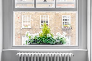 A window with a view of a brick building, a radiator below, and a window box with white flowers and green foliage.