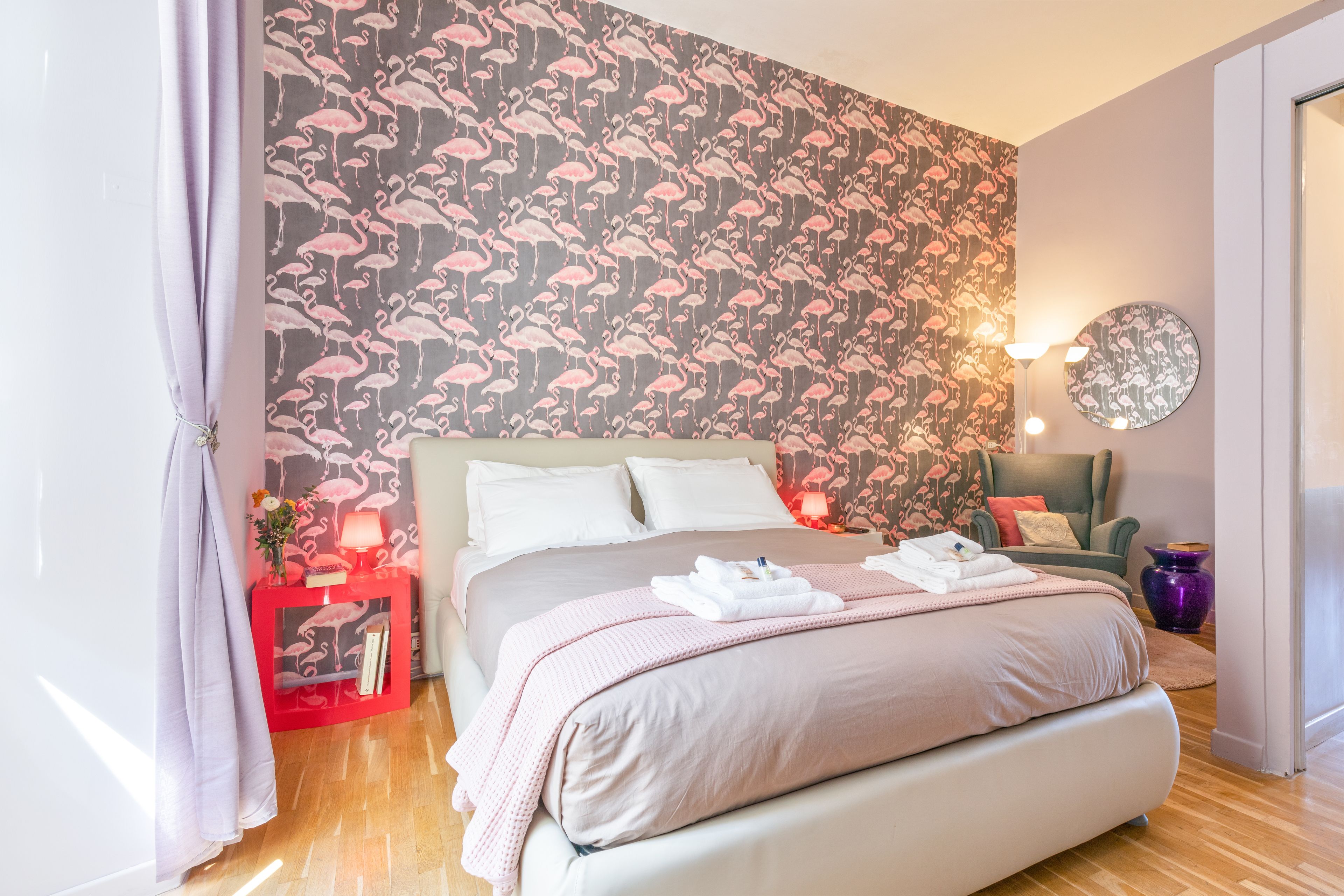 A bedroom with a large bed, a red nightstand, a round mirror, and a wall with a flamingo pattern.