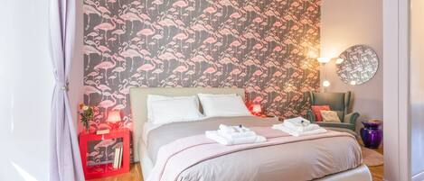 A bedroom with a large bed, a red nightstand, a round mirror, and a wall with a flamingo pattern.