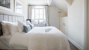 Ein Schlafzimmer mit einem großen Bett, einem Fenster mit Vorhängen, einem Heizkörper und einem Nachttisch mit einer Lampe.