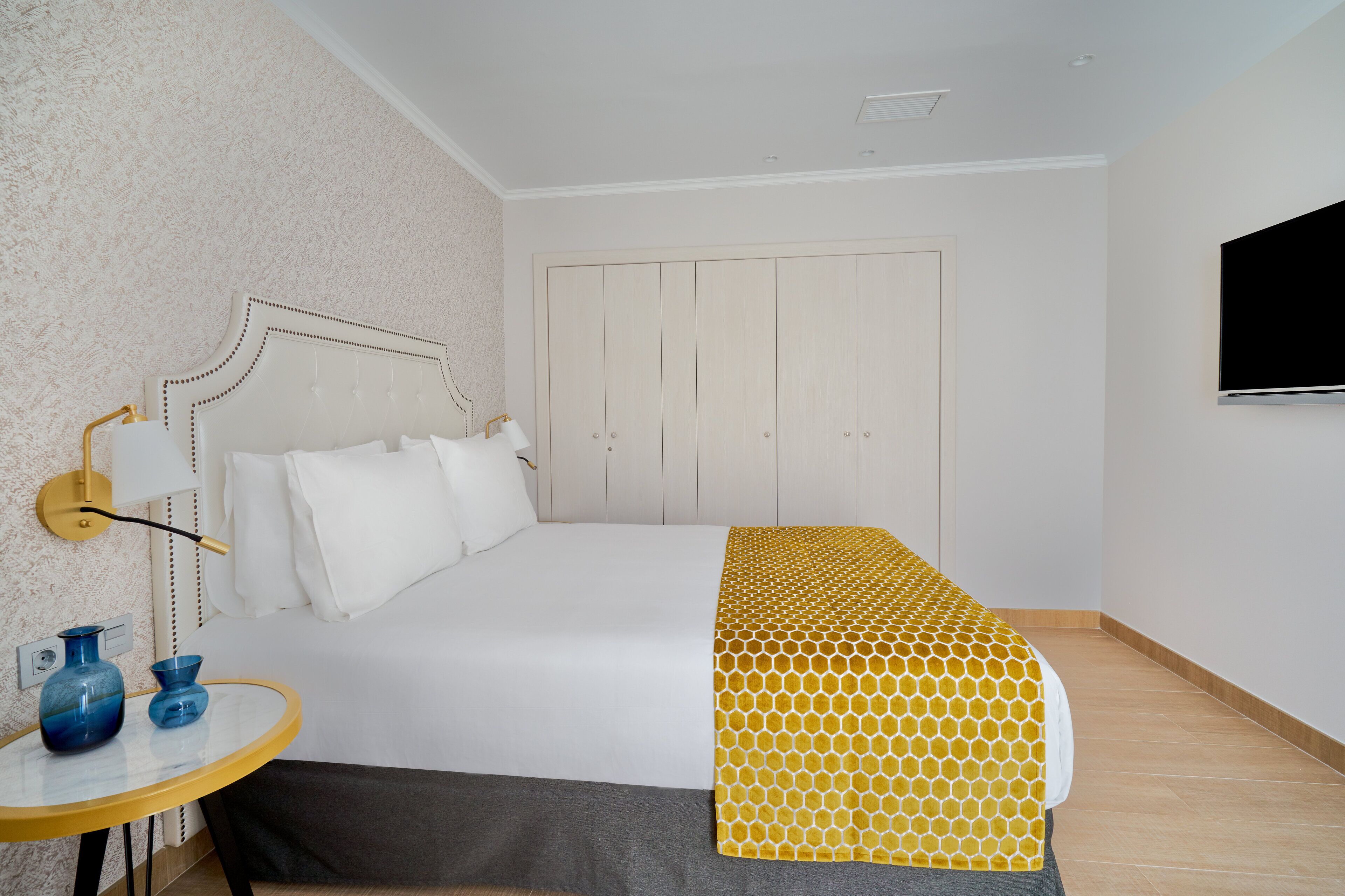 Una habitación de hotel con cama, una mesita de noche con dos jarrones, un televisor montado en la pared y un armario.
