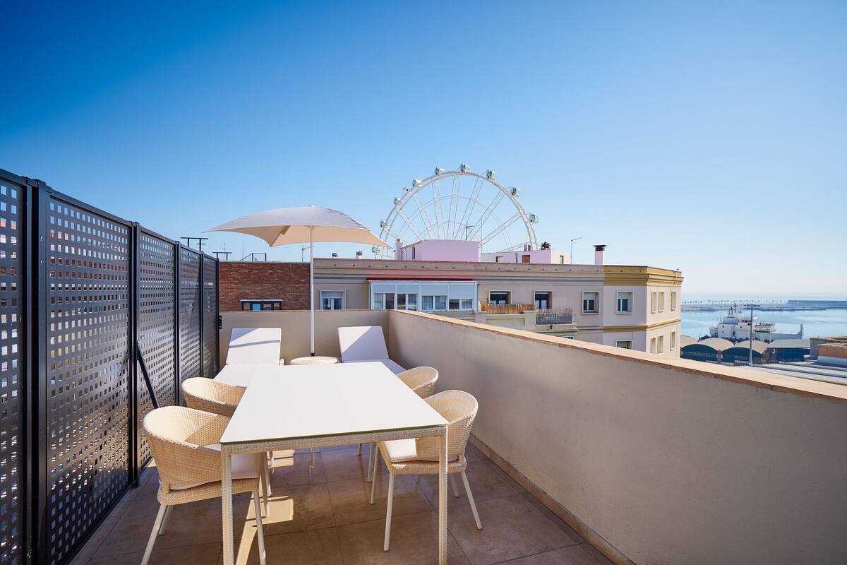 Una terraza en la azotea con una mesa y sillas, una noria al fondo y vista al mar.