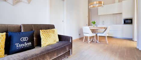 A modern living room with a dark brown sofa, a dining table with chairs, and a kitchen area with white cabinets and a microwave.