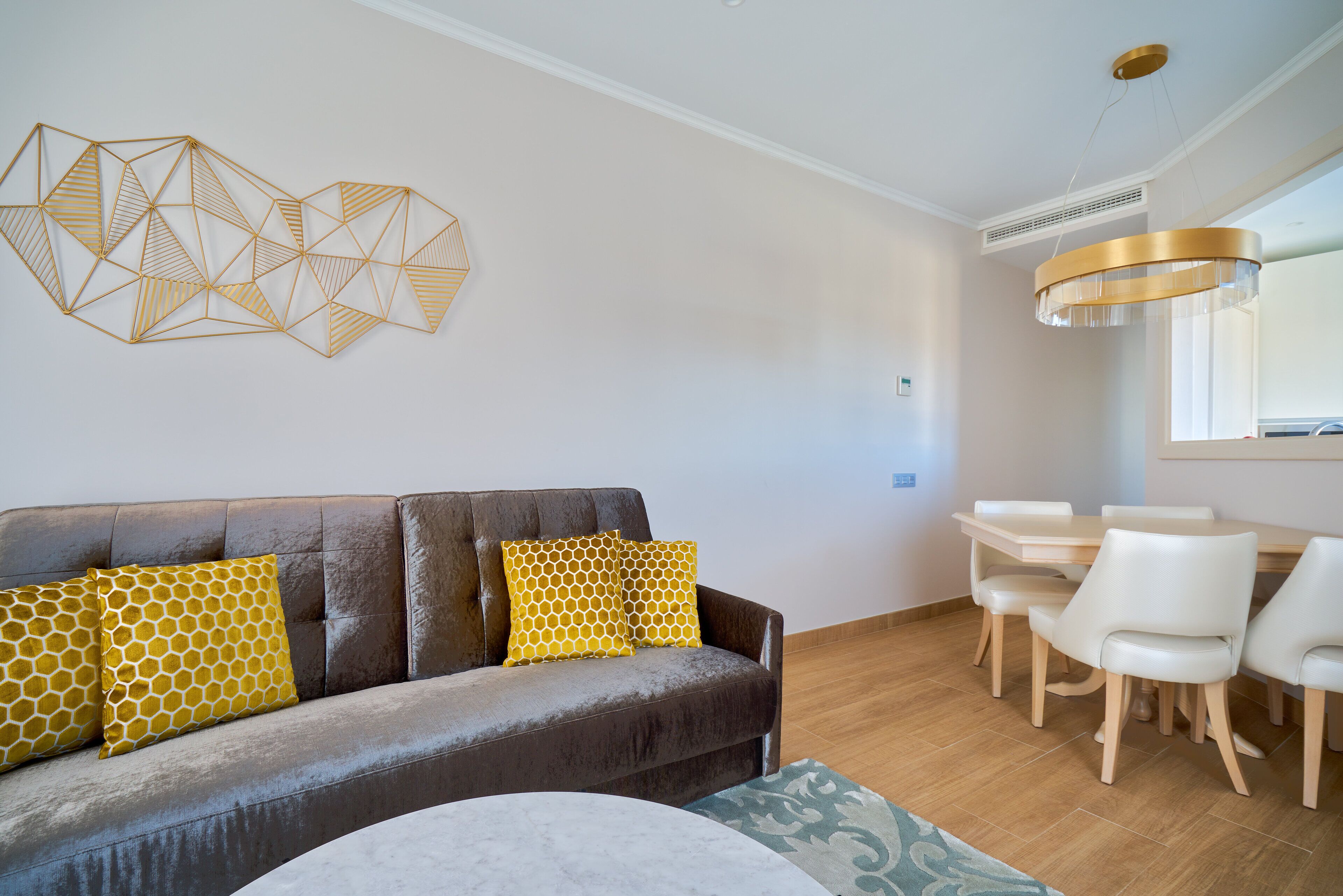 Een moderne woonkamer met een bank, gele kussens met een patroon, een eettafel met stoelen, en een abstract kunstwerk aan de muur.