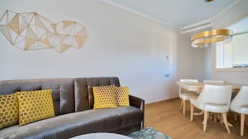 Een moderne woonkamer met een bank, gele kussens met een patroon, een eettafel met stoelen, en een abstract kunstwerk aan de muur.