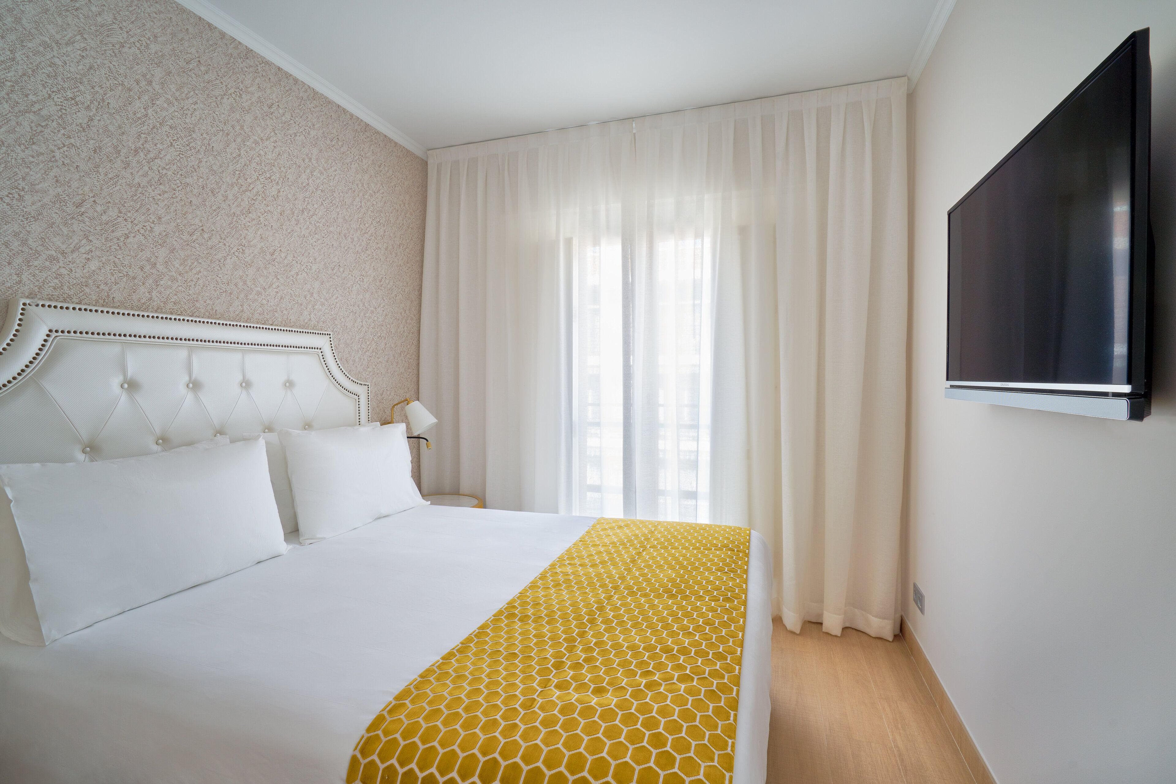 Une chambre d’hôtel avec un grand lit, un téléviseur fixé au mur et une fenêtre avec des rideaux légers.