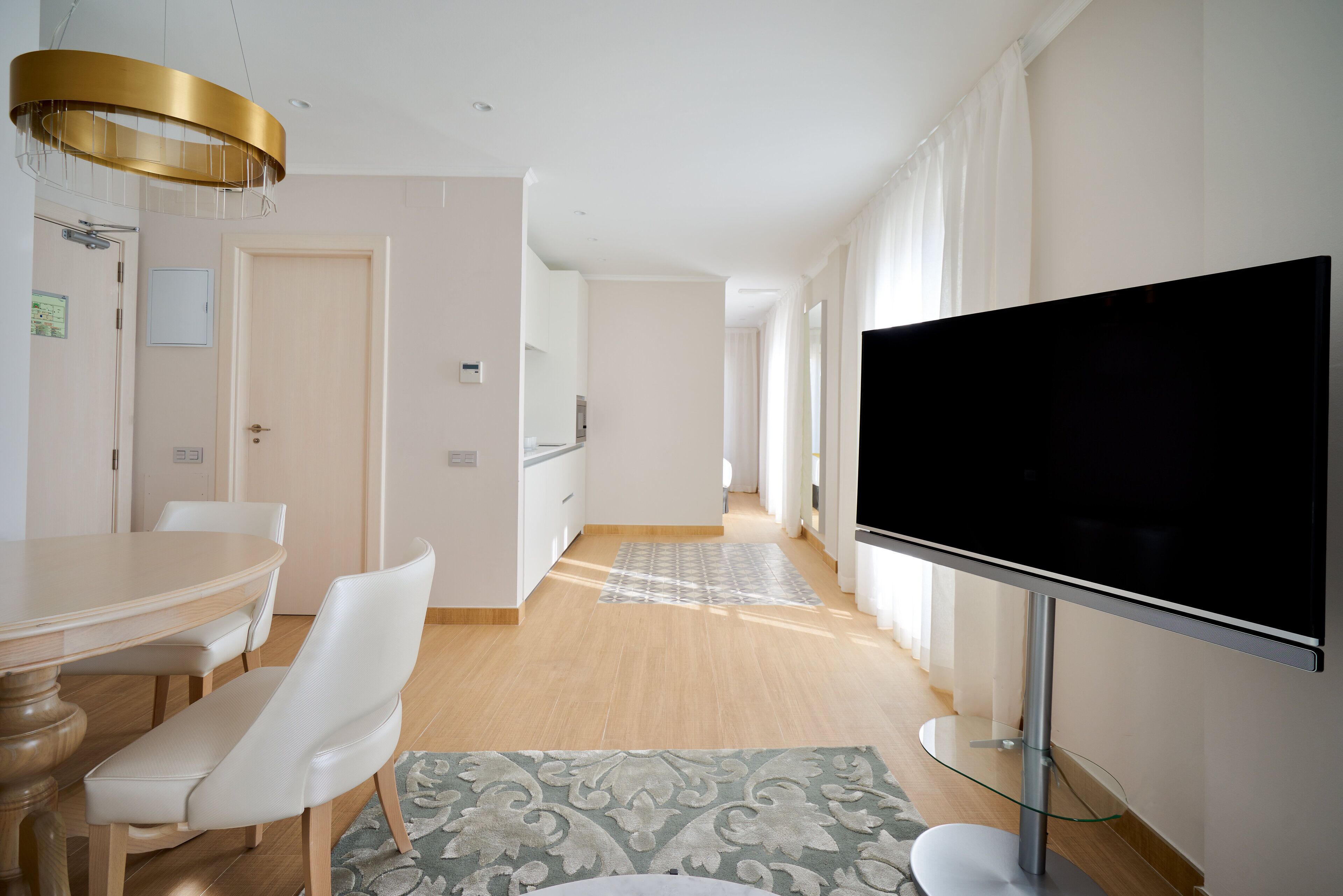 A modern living room with a flat-screen TV, a dining area with a wooden table and white chairs, and a hallway leading to another room.