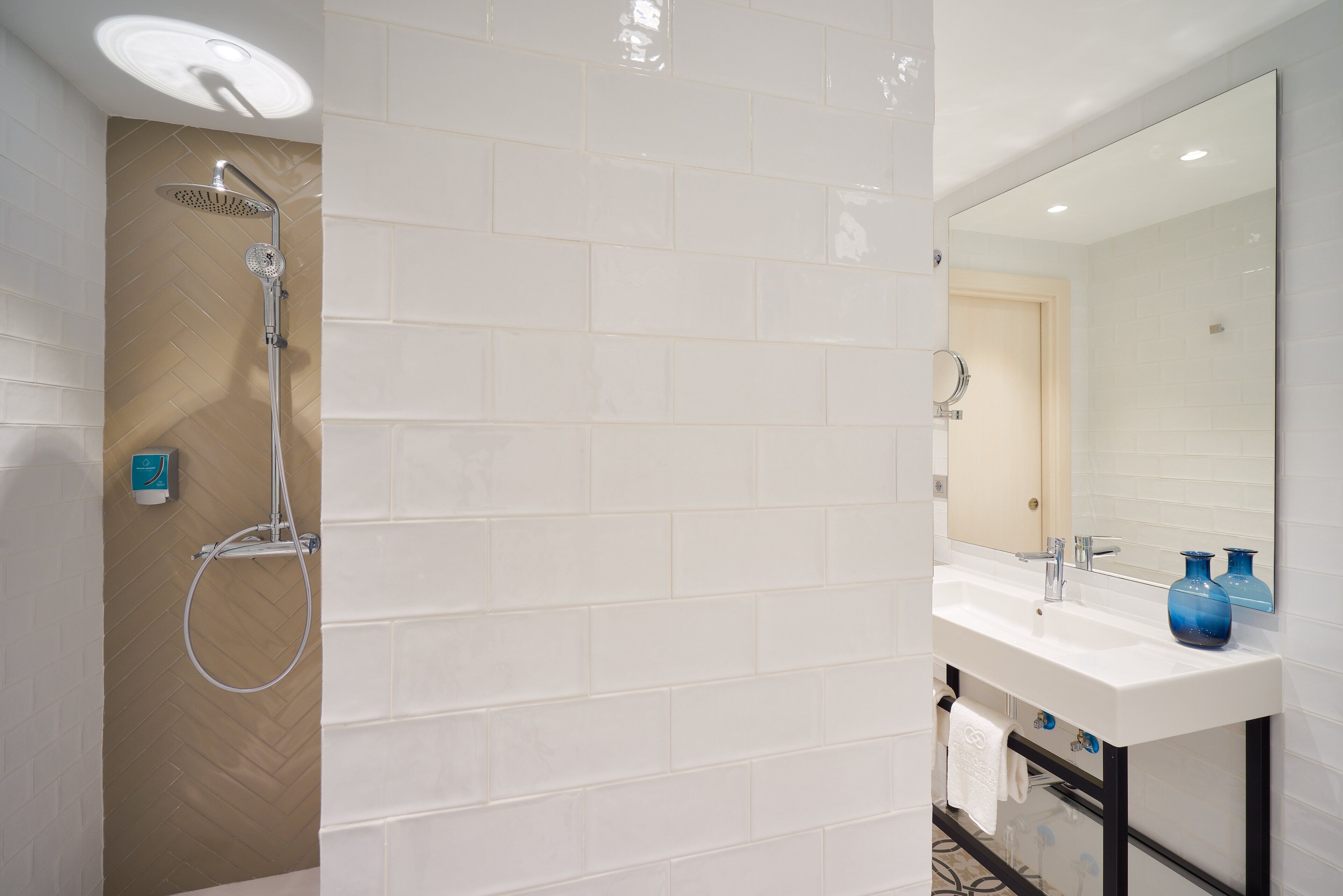 A modern bathroom with a shower, white tiles, a vanity with a mirror, and a blue vase.