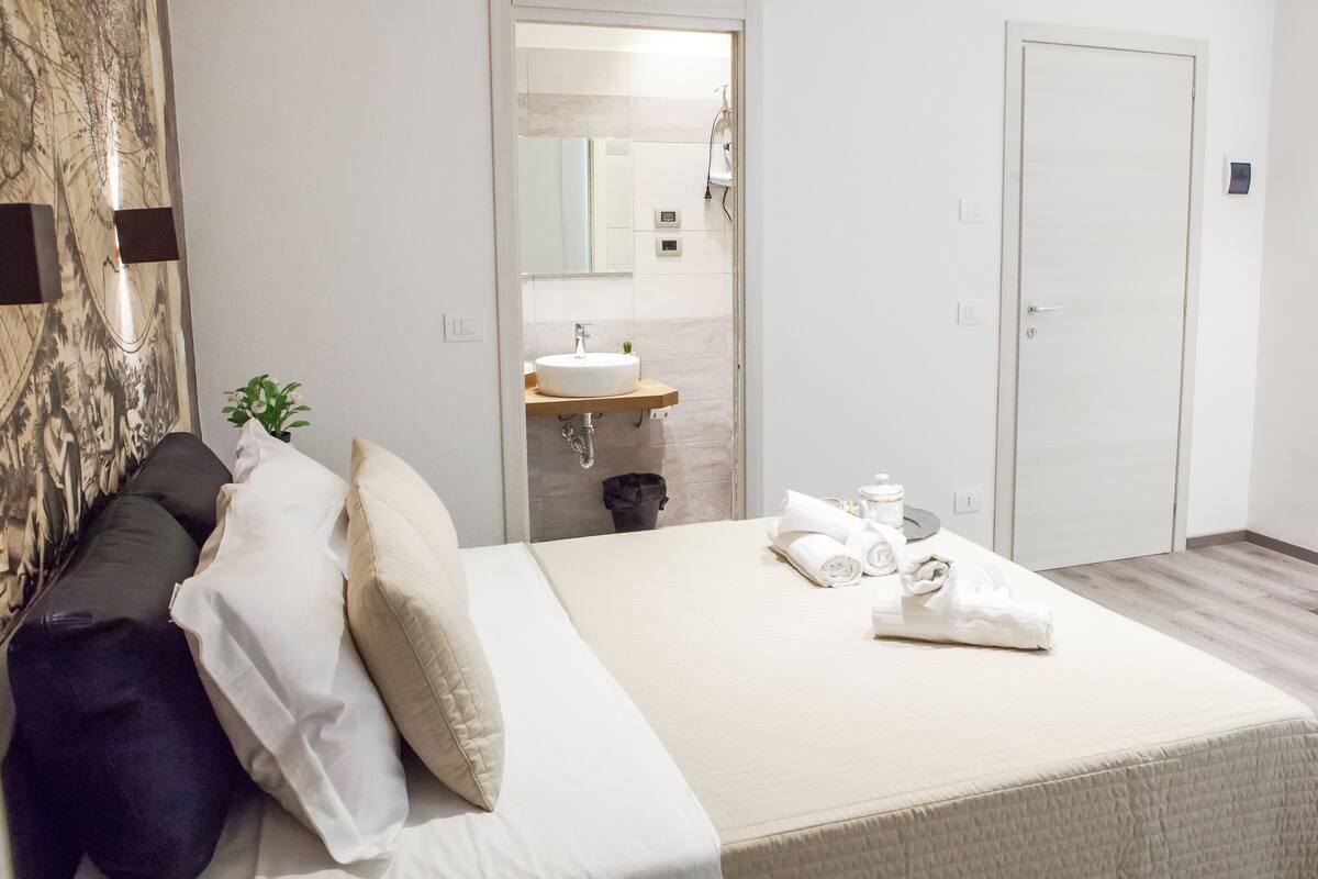 Ein Hotelzimmer mit Bett, Kissen, Nachttisch, Waschbecken und Spiegel.