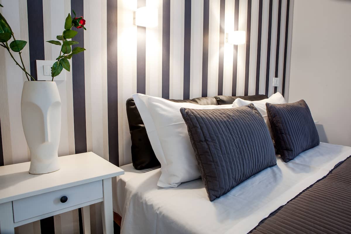 Ein ordentlich gemachtes Bett mit weißen und dunklen Kissen, ein weißer Nachttisch mit einer Vase und einer Rose, sowie eine gestreifte Wand im Hintergrund.