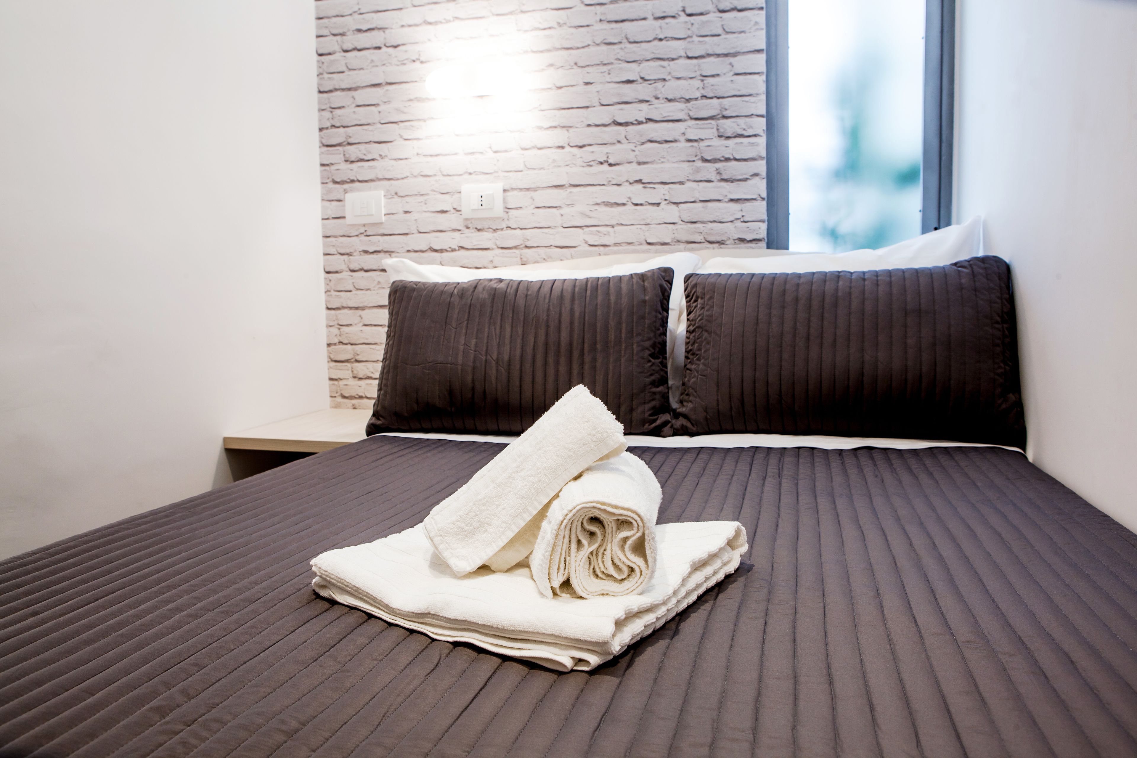 A neatly made bed with a folded towel and two pillows, a brick-patterned wall, and a window with a view.