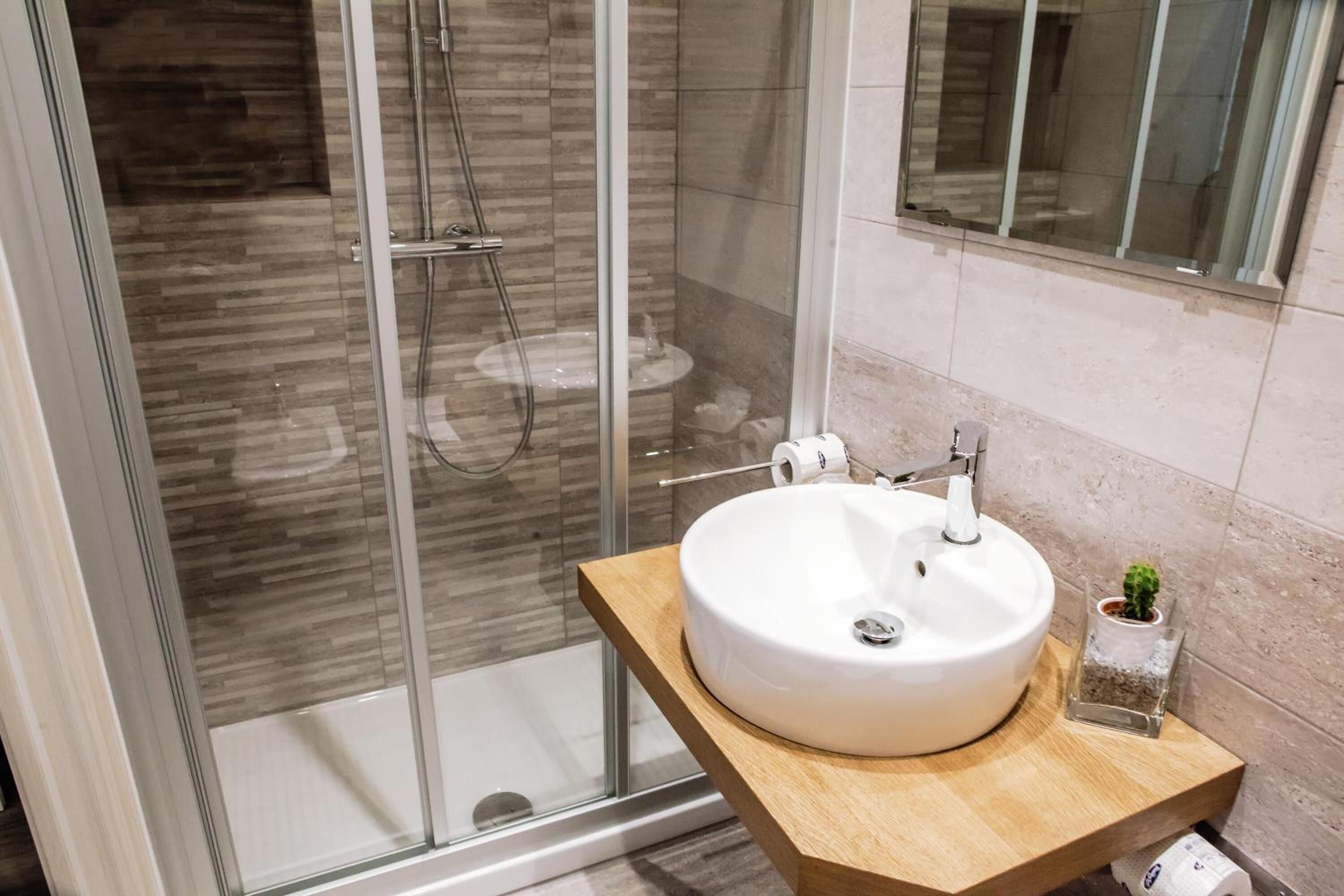 Un baño moderno con cabina de ducha de vidrio, un lavamanos blanco con encimera de madera y una pequeña planta en maceta.