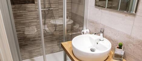 Un baño moderno con cabina de ducha de vidrio, un lavamanos blanco con encimera de madera y una pequeña planta en maceta.