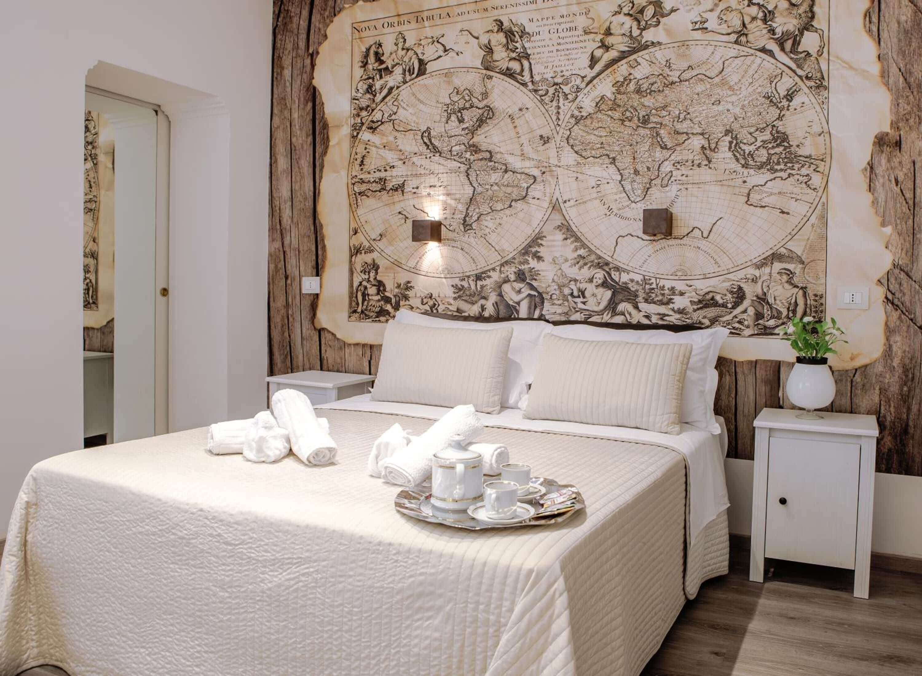A hotel room with a large antique map on the wall, a neatly made bed with white linens, and a tray with tea cups and a teapot.