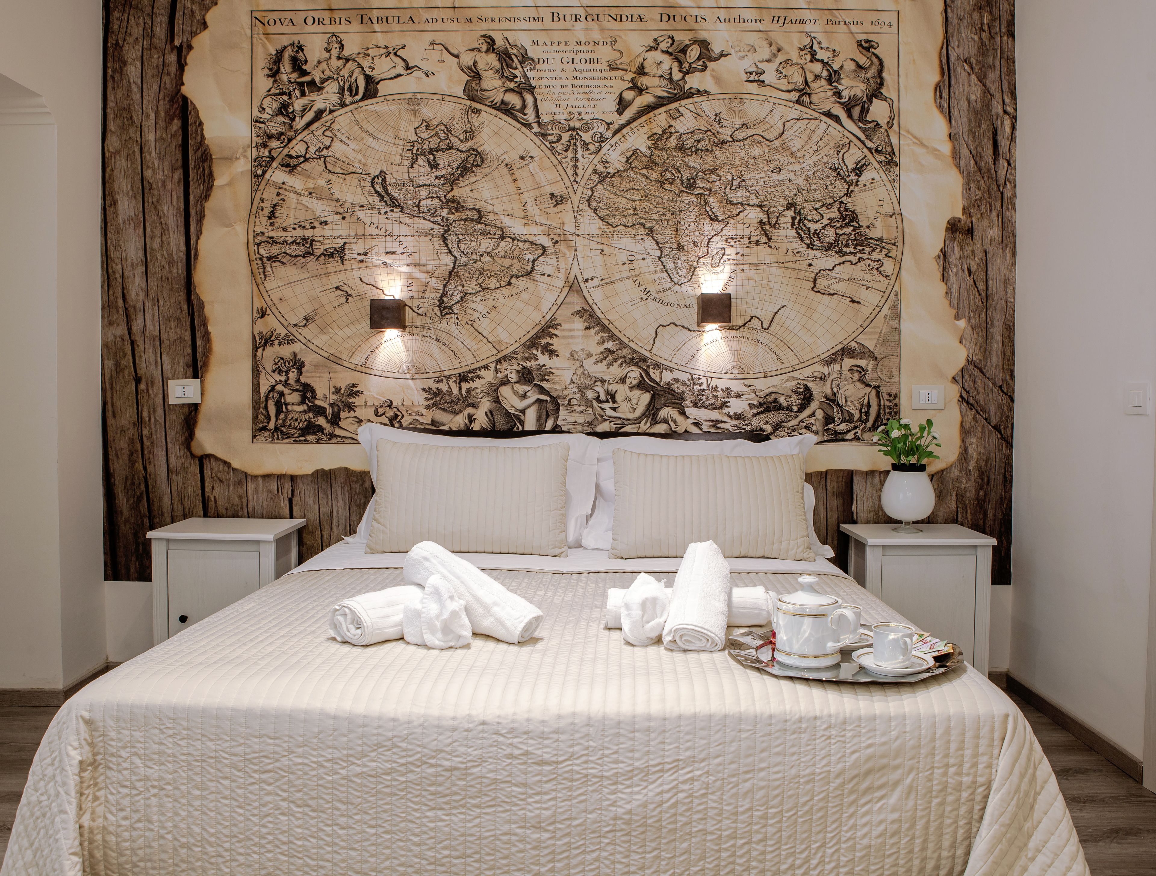Une chambre d’hôtel avec une grande fresque représentant une carte géographique au mur, un lit bien fait avec des serviettes, et un plateau avec une théière et des tasses.