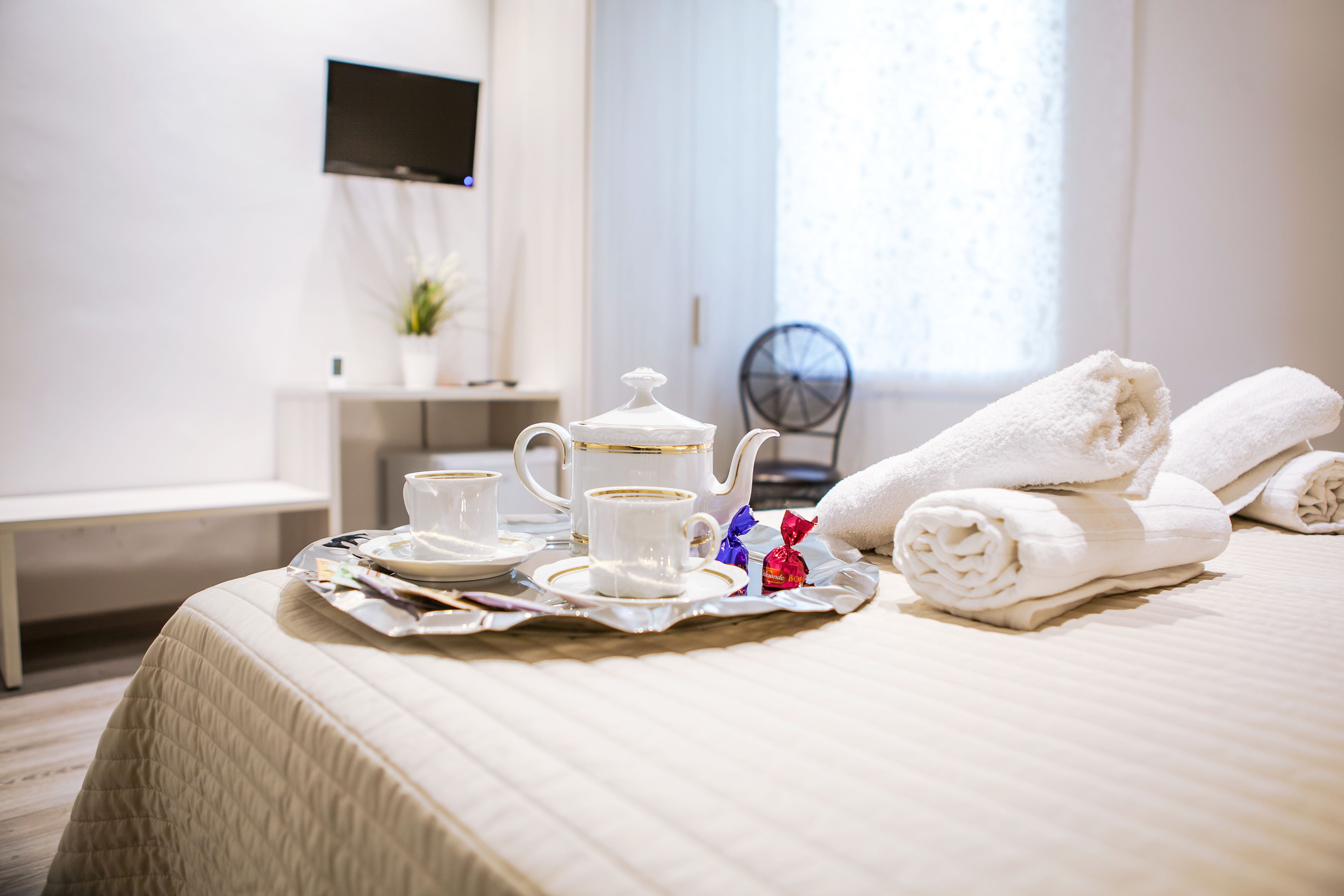 A neatly made bed with a white duvet, a tray with a teapot, cups, and a small red gift. There are towels and a chair in the background.