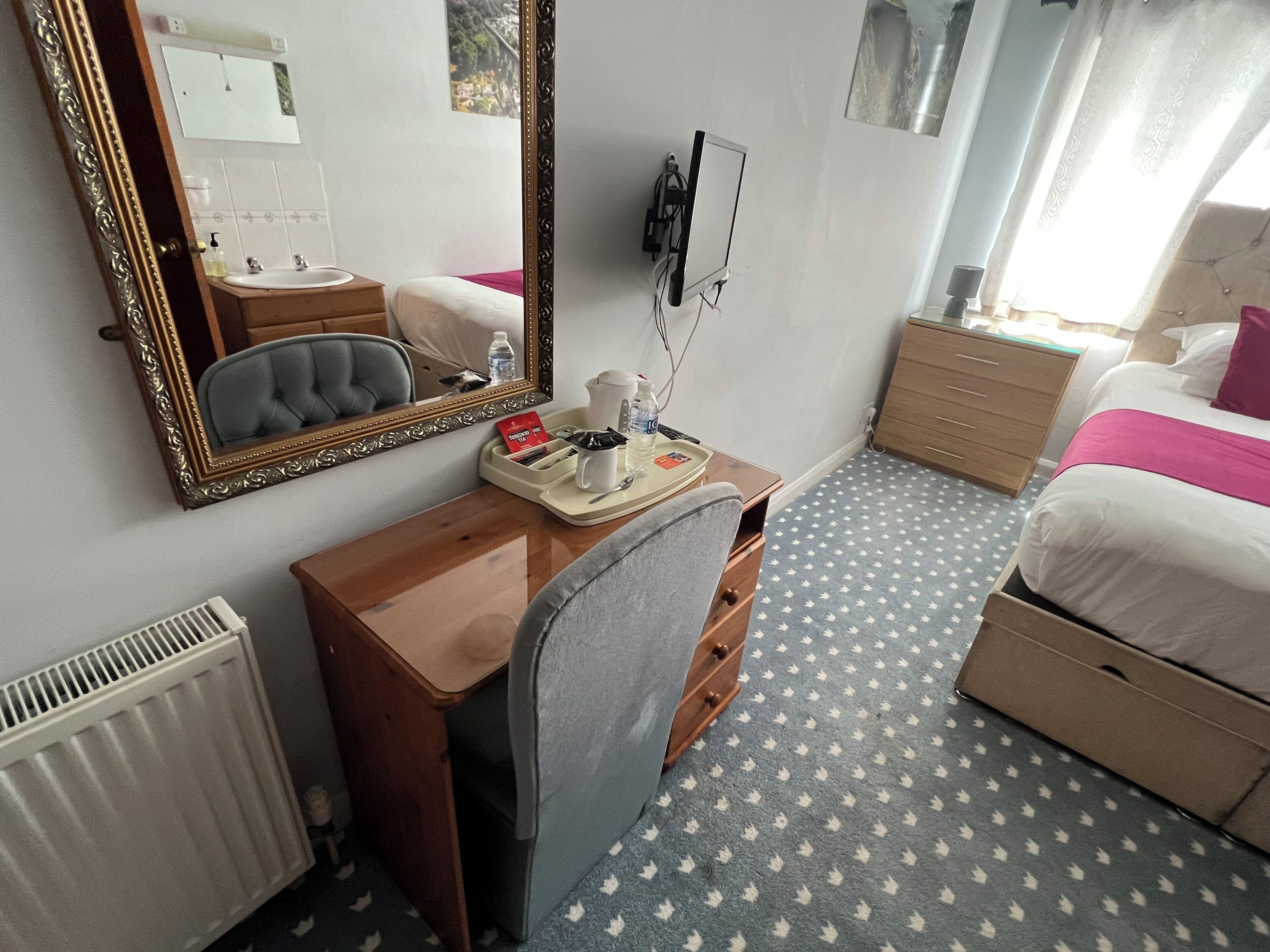 Habitación de hotel con un escritorio de madera, un espejo grande, una cama con ropa de cama blanca y rosa, un radiador y una alfombra estampada.