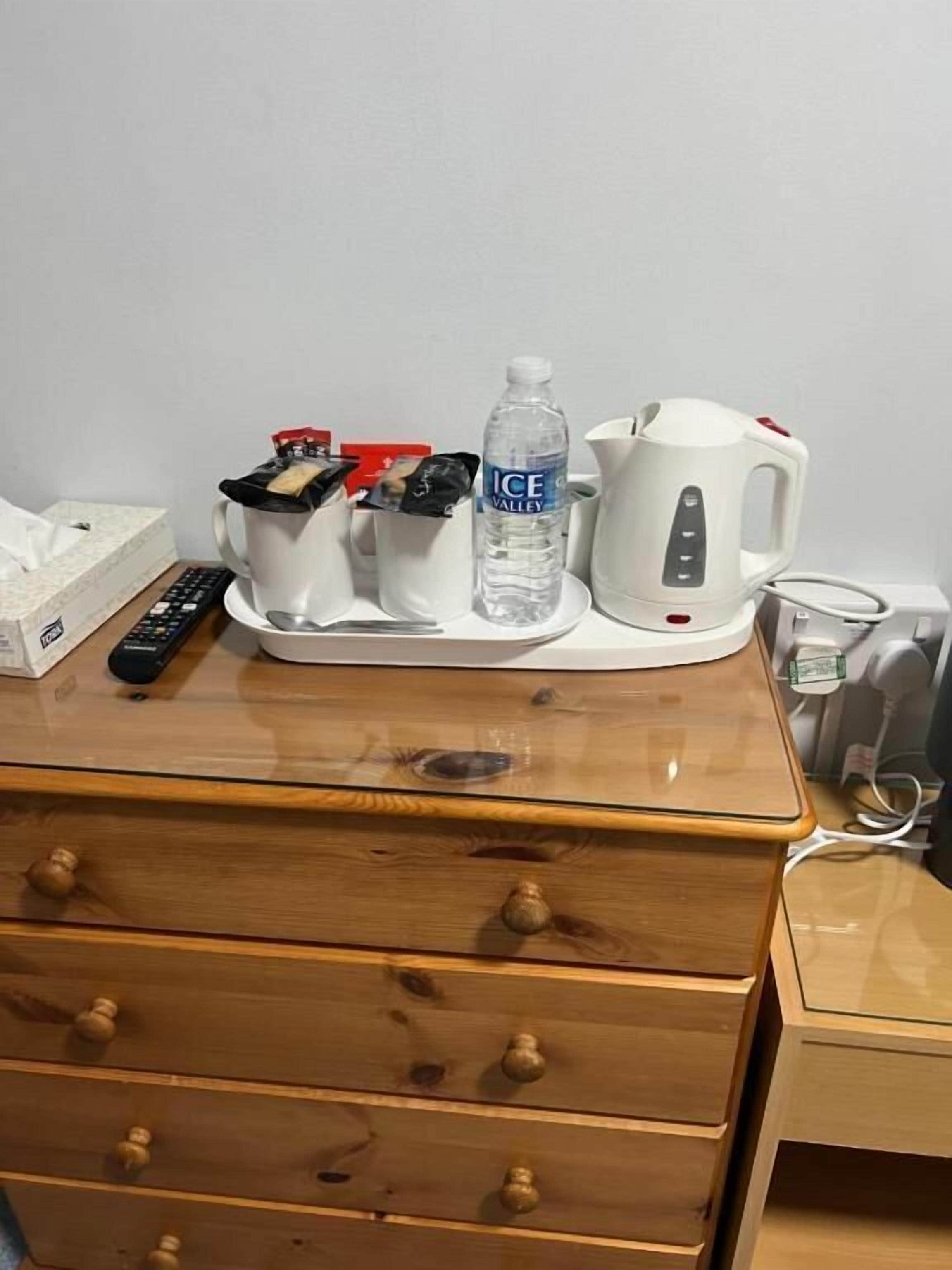 Un comò in legno con cassetti, un telecomando, una scatola di fazzoletti, una tazza bianca, una bottiglia d'acqua e un bollitore elettrico bianco sopra.