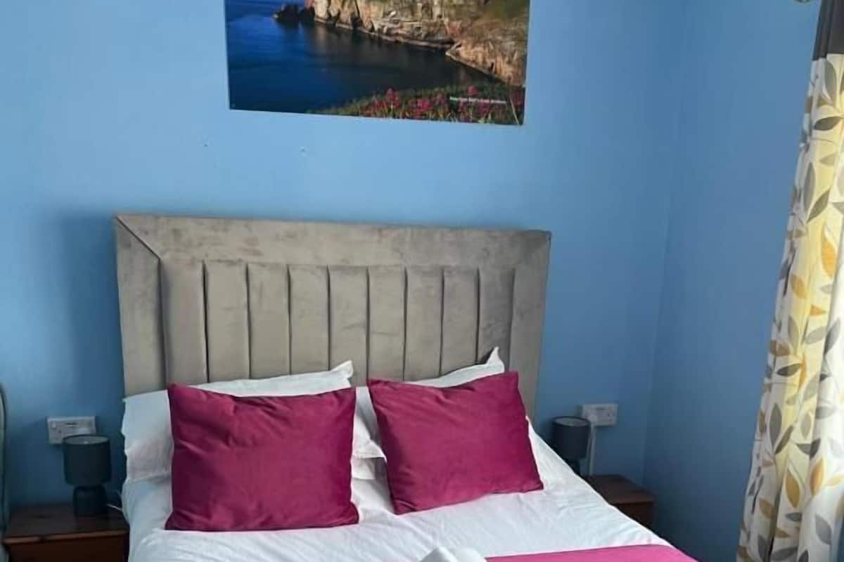 A neatly made bed with a headboard, two pillows, a rolled towel, and a painting of a coastal scene above it.